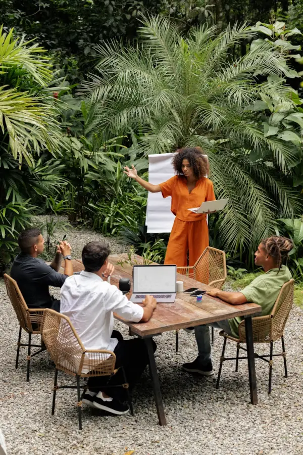 Une réunion d’équipe en plein air autour d’une table en bois, une collègue présentant devant un paperboard, au milieu des palmiers et de la végétation tropicale