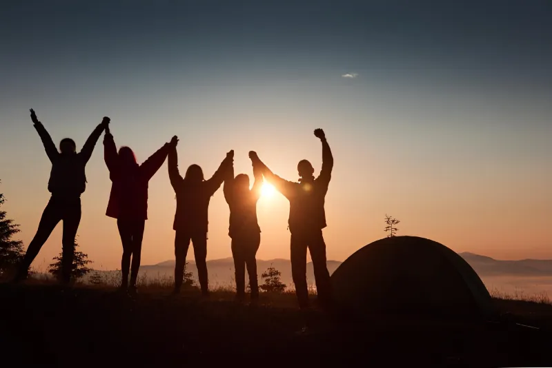 Cinq personnes bras dessus bras dessous sur une crête au coucher du soleil, en contre-jour, une tente dressée à côté d’elles
