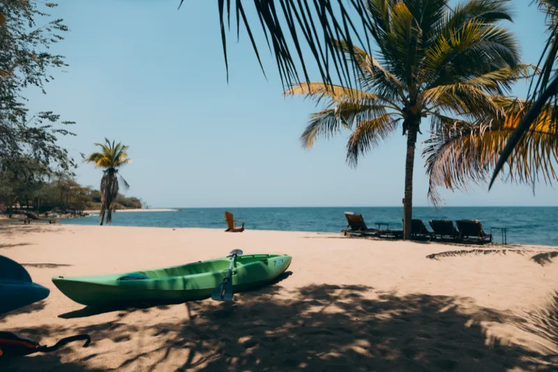 A green kayak and paddle resting on the sand in the shade of coconut palms, sun loungers and the blue water of Lake Tanganyika beyond