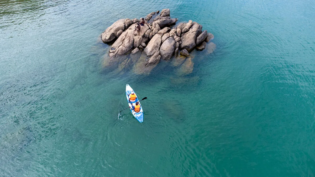 Two people paddling a kayak in turquoise water beside a rock outcrop, seen from above