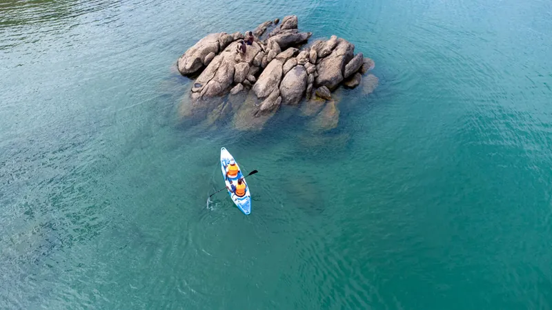 Two people paddling a kayak in turquoise water beside a rock outcrop, seen from above