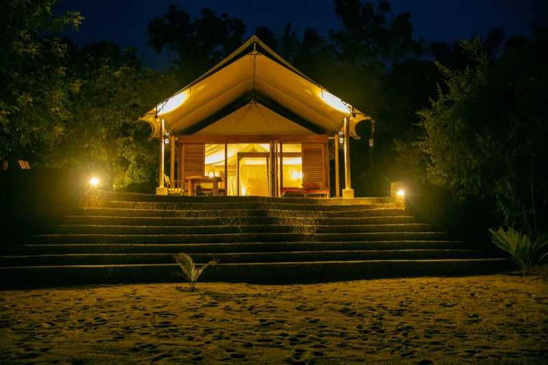 Une tente de safari de luxe éclairée au-dessus des marches de la plage, sous un ciel nocturne d’un bleu profond