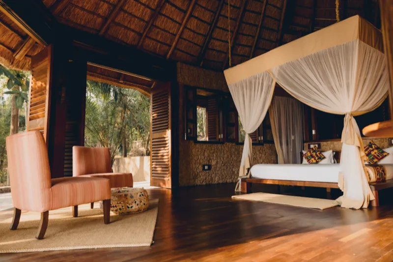 L’intérieur de la villa Obsidian : un lit à baldaquin voilé de moustiquaire blanche sous un haut toit de chaume, deux fauteuils corail sur un tapis tressé et des portes à persiennes ouvertes sur les palmiers