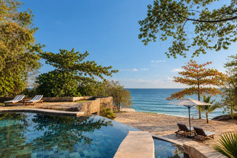 La piscine à débordement sous la villa Obsidian, des transats sur la terrasse en pierre, un parasol blanc sur la plage privée et le lac Tanganyika sous un ciel bleu