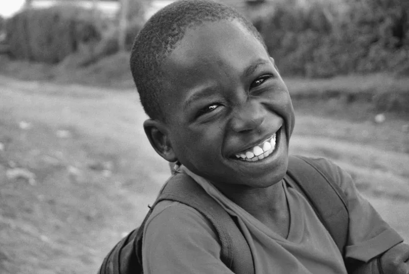 Un enfant portant un sac à dos, riant par-dessus son épaule sur un chemin de terre, des arbres derrière lui, photographié en noir et blanc