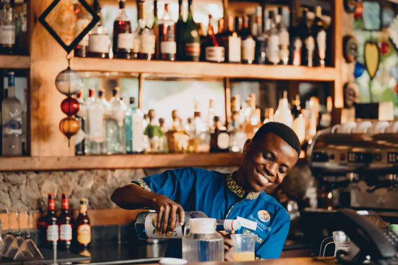 Un barman souriant en uniforme bleu Sunstone verse une boisson au bar, devant des étagères de bouteilles éclairées