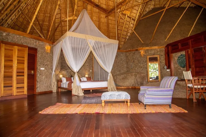 A spacious bedroom with a canopied bed, hardwood floor and stone walls under a thatched ceiling