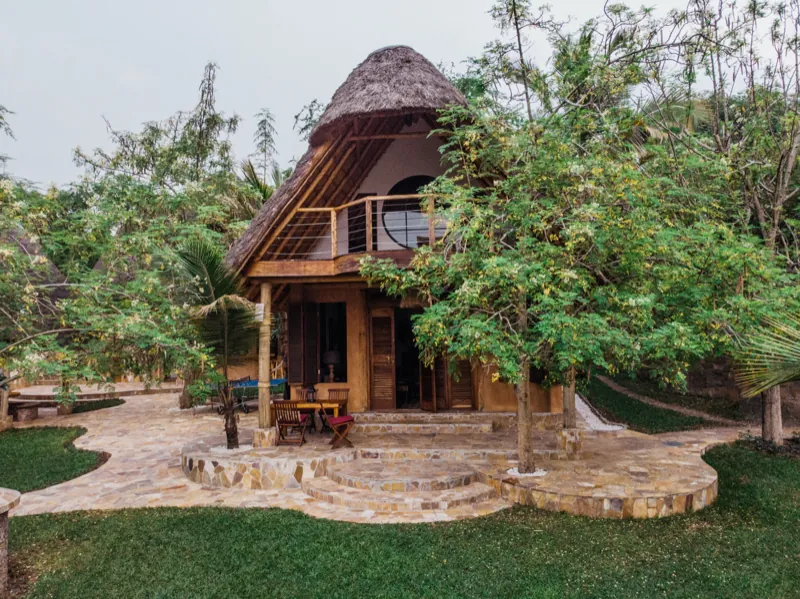 A conical thatched building with an upper balcony and a dining table on a curved flagstone terrace