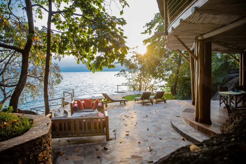 Une terrasse dallée ombragée au bord de l’eau, avec lits de repos et transats à coussins, à côté d’une terrasse en bois ouverte sur le lac Tanganyika au coucher du soleil