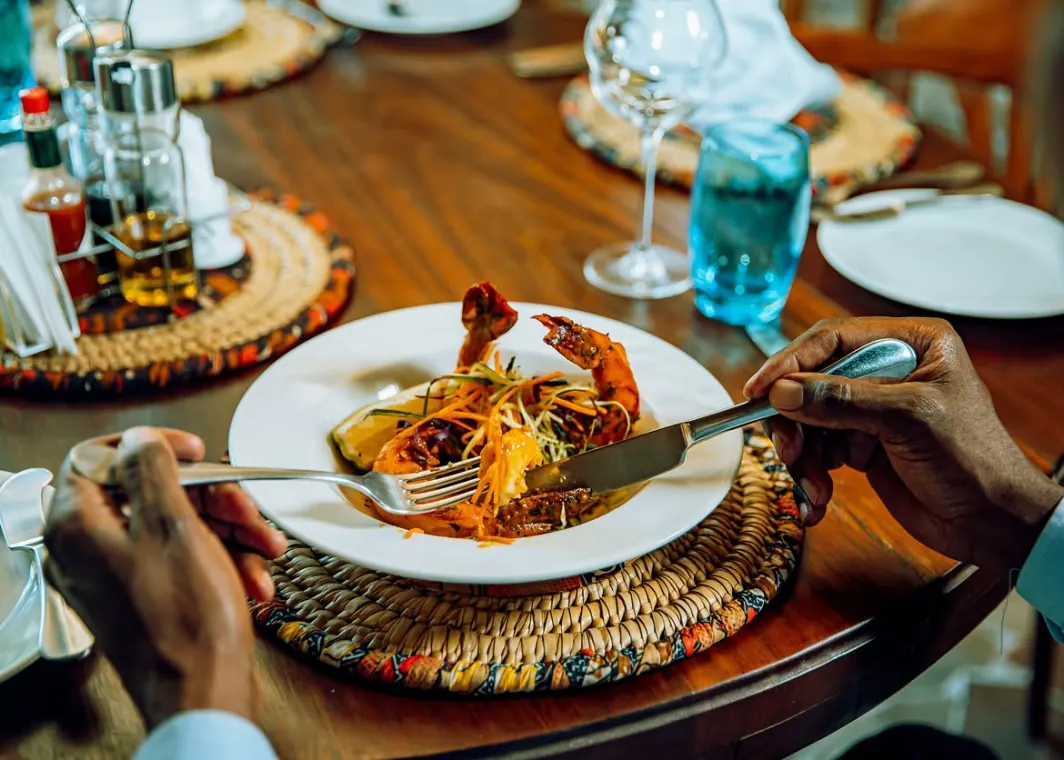 A guest cutting into a plated prawn dish at a laid table, woven mats and glassware around it