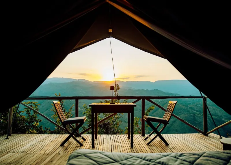 La vue depuis l’intérieur d’une tente perchée : deux chaises pliantes et une table sur le deck, les vallées verdoyantes au-delà et le soleil qui descend derrière elles