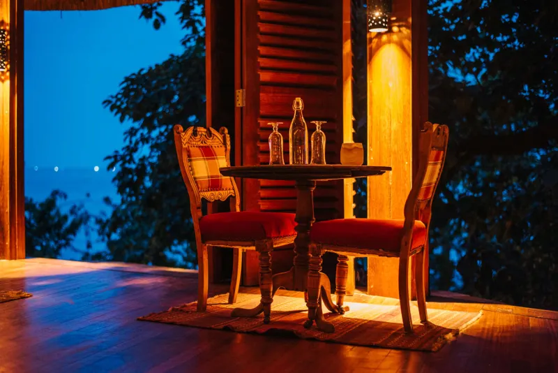 Deux chaises sculptées autour d’une petite table sur une terrasse en bois, éclairées aux lampes devant le bleu du lac à la tombée de la nuit