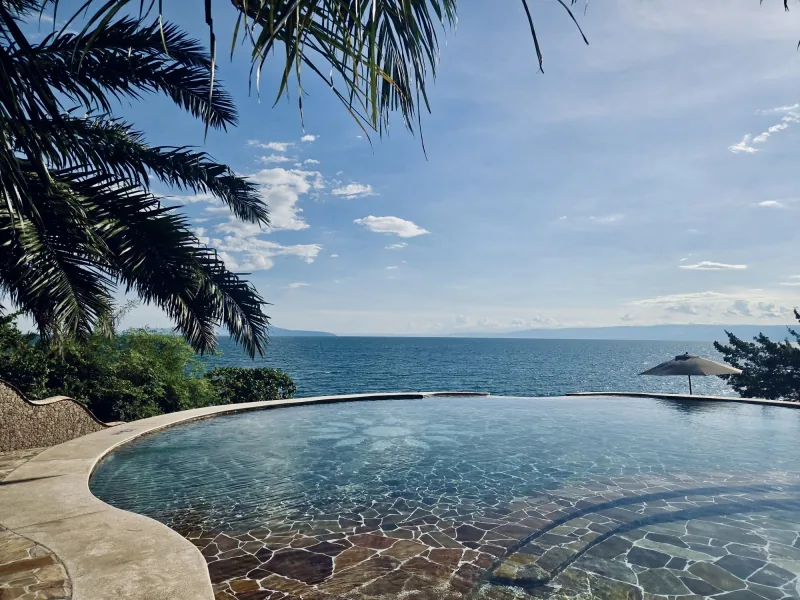 La grande piscine à débordement, ouverte sur le bleu profond du lac Tanganyika et les collines au loin, encadrée par un palmier et un parasol sur la terrasse