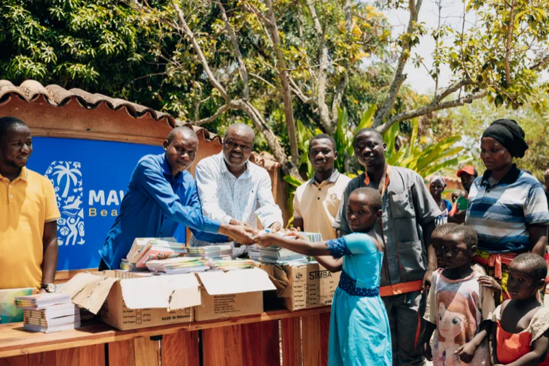 Une fillette en robe bleue tend la main au-dessus d’une table couverte de cahiers pour en recevoir un d’un groupe d’hommes souriants, d’autres enfants et des voisins attendant à ses côtés sous les arbres