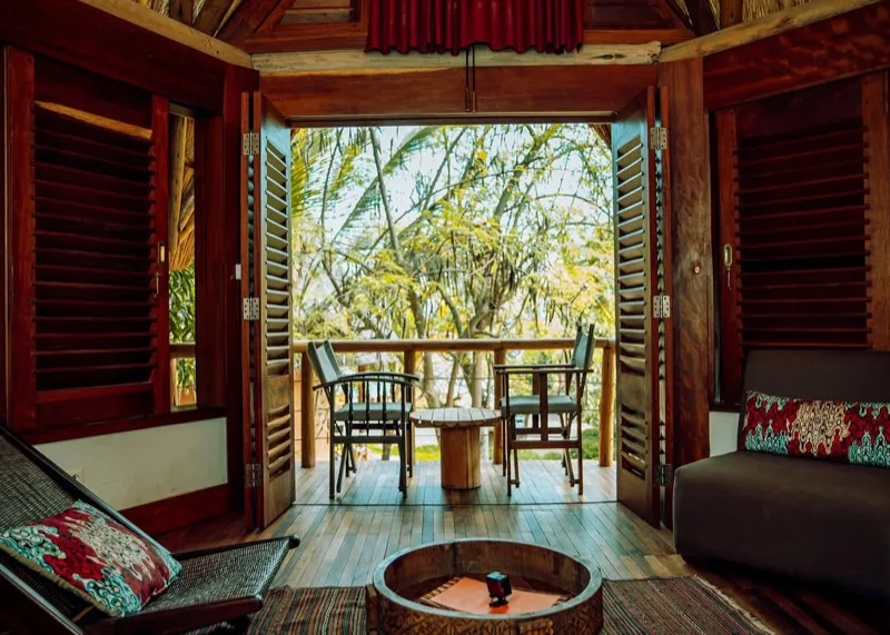 Une chambre de Sunstone Beach dont les portes à persiennes s’ouvrent sur un balcon privé avec table et chaises, le jardin au-delà