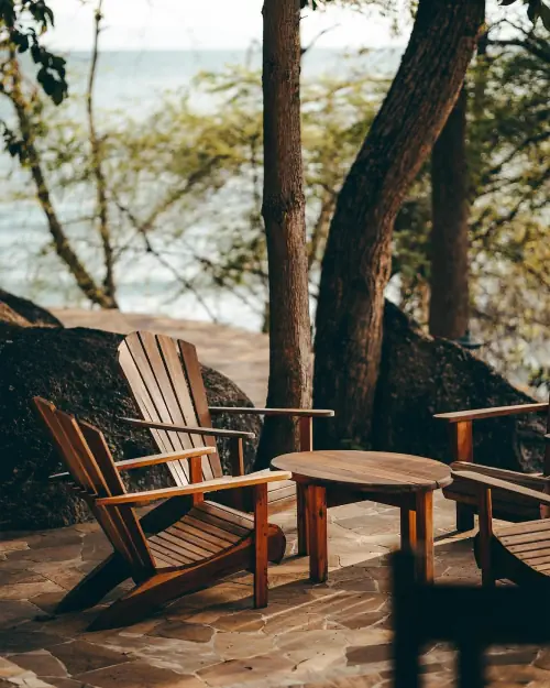 Two wooden armchairs and a round table on flagstones under the trees, at the water’s edge