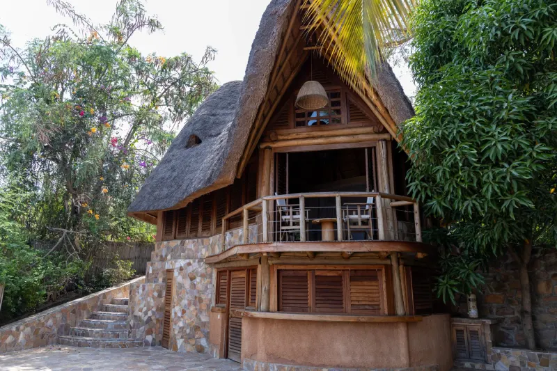 Des marches de pierre montant à la véranda du bungalow Rubis, un fauteuil de part et d’autre des portes à persiennes, sous un large toit de chaume
