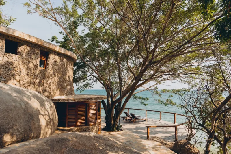 A shaded stone terrace at the water’s edge, cushioned daybeds and loungers facing Lake Tanganyika with the low sun through the trees