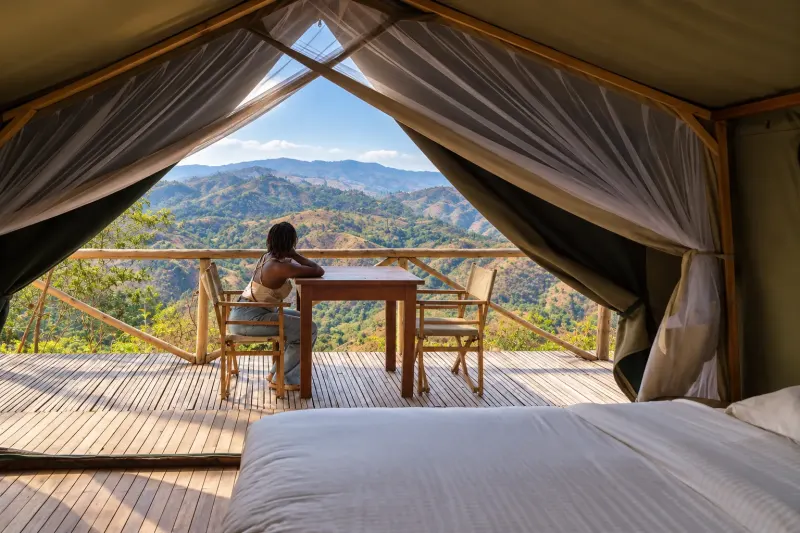 La vue depuis une tente perchée : deux chaises pliantes et une table sur la terrasse, les vallées verdoyantes au-delà et le soleil qui se couche derrière