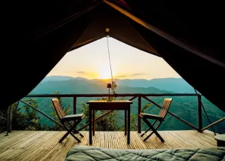 La vue depuis une tente perchée : deux chaises pliantes et une table sur la terrasse, les vallées verdoyantes au-delà et le soleil qui se couche derrière